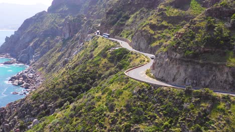 Eine-Luftaufnahme-Eines-Konvois-Von-Bussen,-Die-Auf-Einer-Gefährlichen-Schmalen-Bergstraße-Entlang-Der-Ocean-Chapmans-Peak-Road-In-Der-Nähe-Von-Kapstadt-Südafrika-Fahren