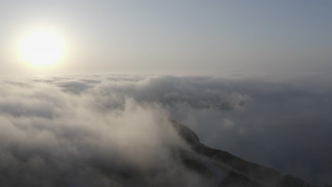 Epische-Vogelperspektive-Auf-Die-Meeresküste-Bei-Sonnenuntergang-Mit-Dichten-Nebelwolken,-Die-über-Die-Hügel-Ziehen-Und-Sie-Vollständig-Bedecken