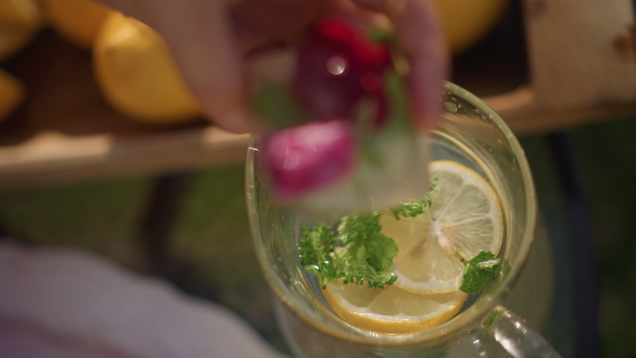 Brocca di limone su tavolo del mercato con menta e petali galleggianti, cassa di limoni del venditore sullo sfondo, una mano che lascia cadere un cubetto di ghiaccio con un petalo di rosa in un bicchiere, bollicine dall'alto intorno alla fetta di agrume, esterno luminoso.