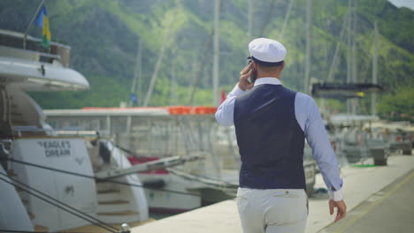 man walking on a dock talking on the phone, by yachts and mountains