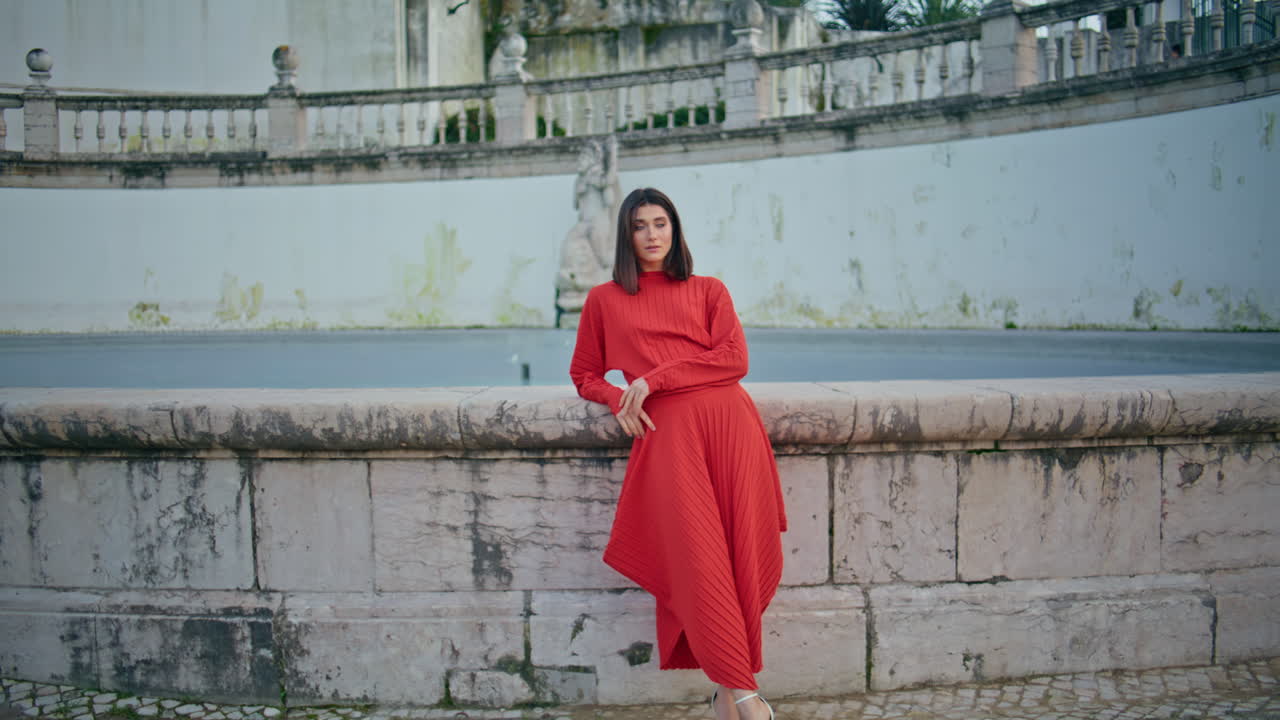Red dress girl looking camera at city square. Chic lady standing town fountain