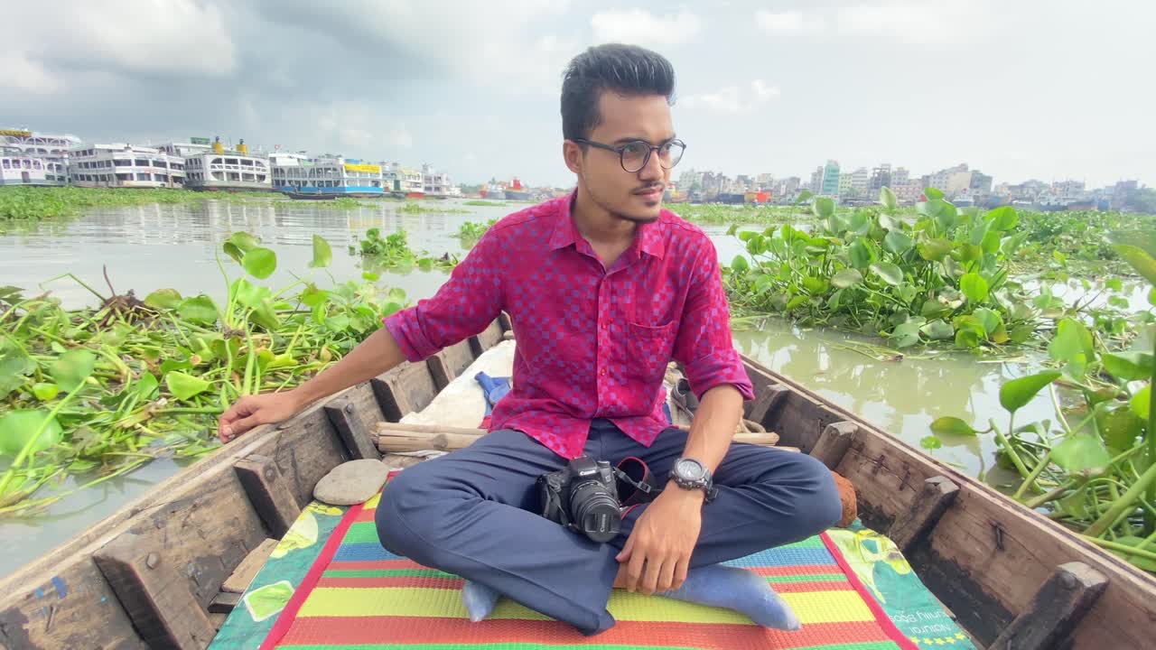 부리강가 강에서 부레옥잠을 지나 떠다니는 무릎에 카메라를 들고 보트에 앉아 있는 방글라데시 남성의 모습