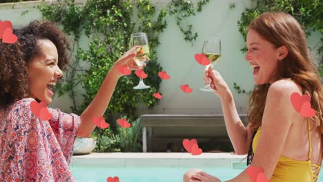 multiple red hearts floating against two diverse female friends toasting drinks by the pool