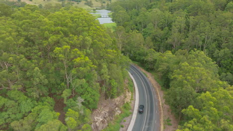 Cyclist-Riding-And-Car-Driving-Through-Scenic-Rural-Road-With-Trees-Alongside-Highway,-4K-Slow-Motion-Drone