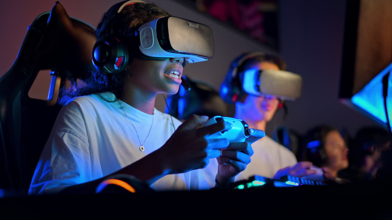 White boy and black girl teens in VR headsets playing video games in video game club with blue illumination using a gamepad