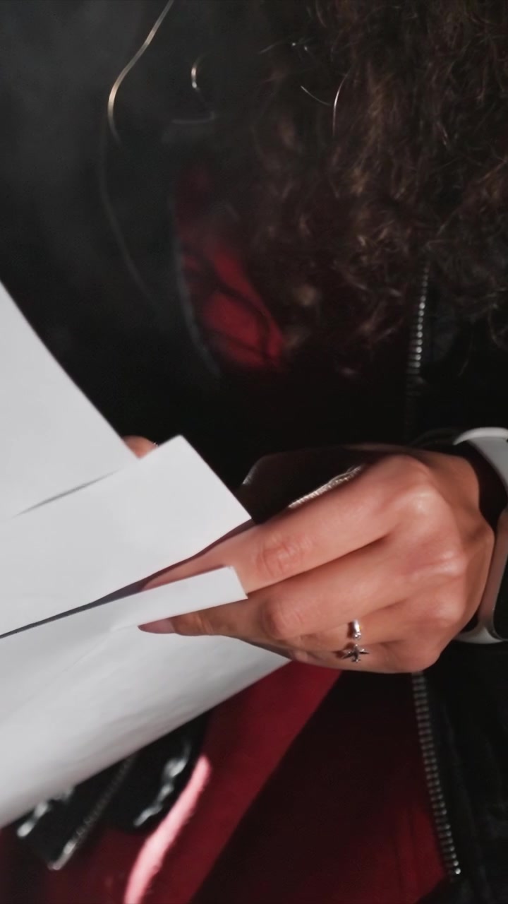 Sujet manipulant une pile de documents sous une lumière tamisée. Des mains baguées feuillettent plusieurs pages de papier. L'arrière-plan sombre met en valeur les actions du sujet sans trop de détails.