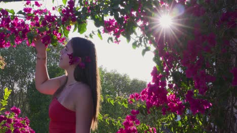 a young model with long black hair looks at the red blossoms of a bush through which the sun's rays peak