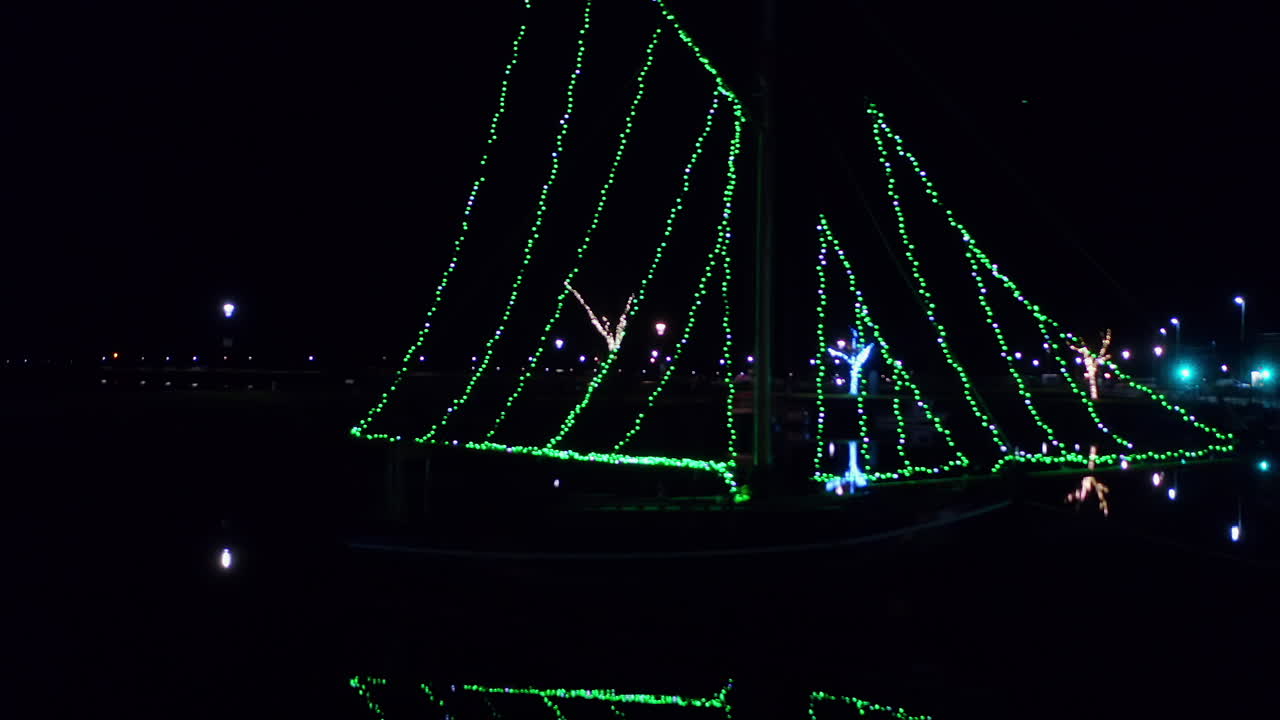 Drone tracking right with Galway Hooker boats strung up with green string lights sparkling in water at night