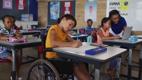 Mixed-race-schoolboy-in-wheelchair,-sitting-in-classroom-making-notes,-teacher-in-background