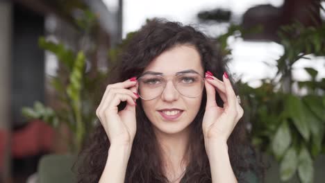 smiling young woman brushes the hair out of her face, looks at the camera, and puts on a pair of glasses