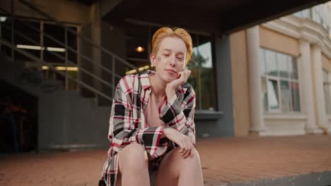 Portrait-of-a-lesbian-girl-with-a-short-haircut-with-sparkles-on-her-face-in-a-pink-checkered-shirt-who-sits-on-a-step-on-the-street,-propping-her-head-up-with-her-palm,-looking-at-the-camera