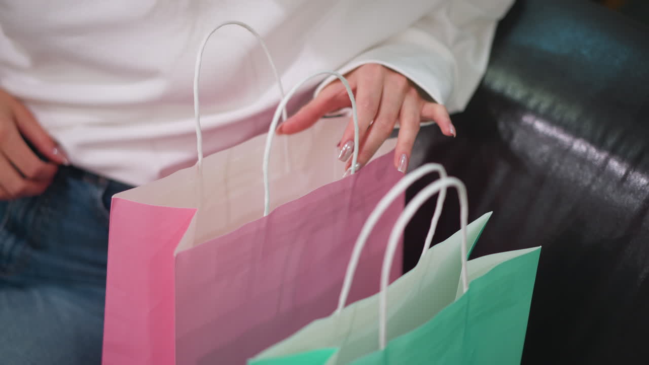 primer plano de una mujer con una camiseta blanca y pantalones vaqueros abriendo bolsas de compras gradualmente en un centro comercial, las uñas pintadas mostrando la elegante bolsa rosa y verde