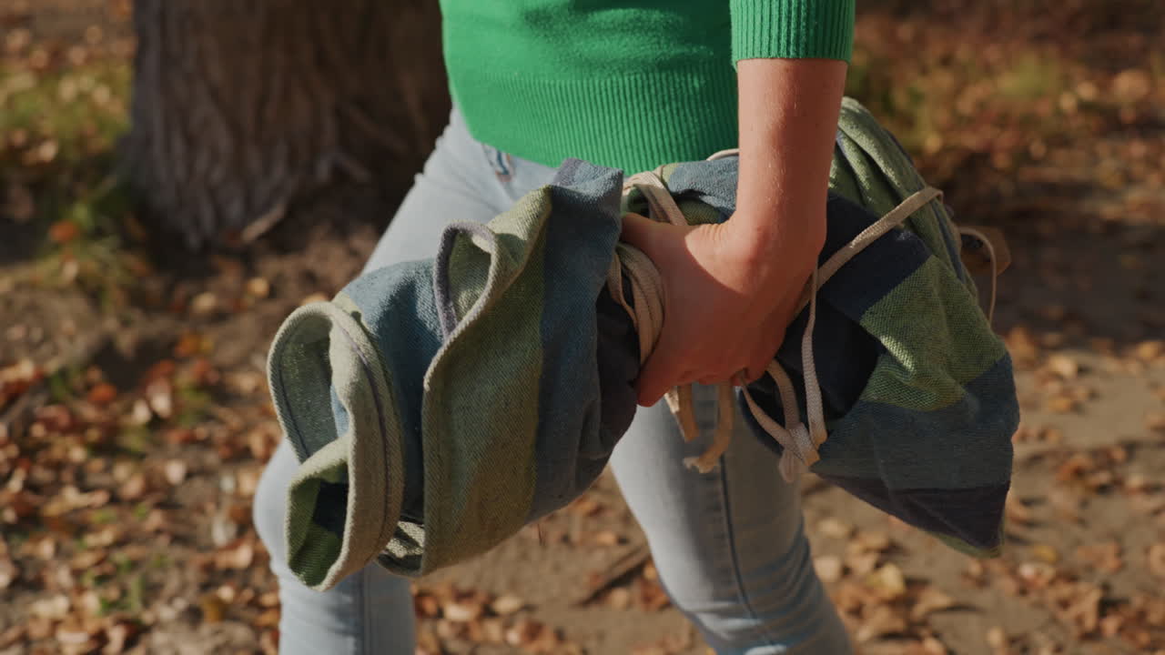Asian woman loading camping blanket from trunk, rolled blanket tied with rope, green sweater and jeans, open car trunk, autumn leaves on ground, clipboard and gear in back, sunlight warmth, calm