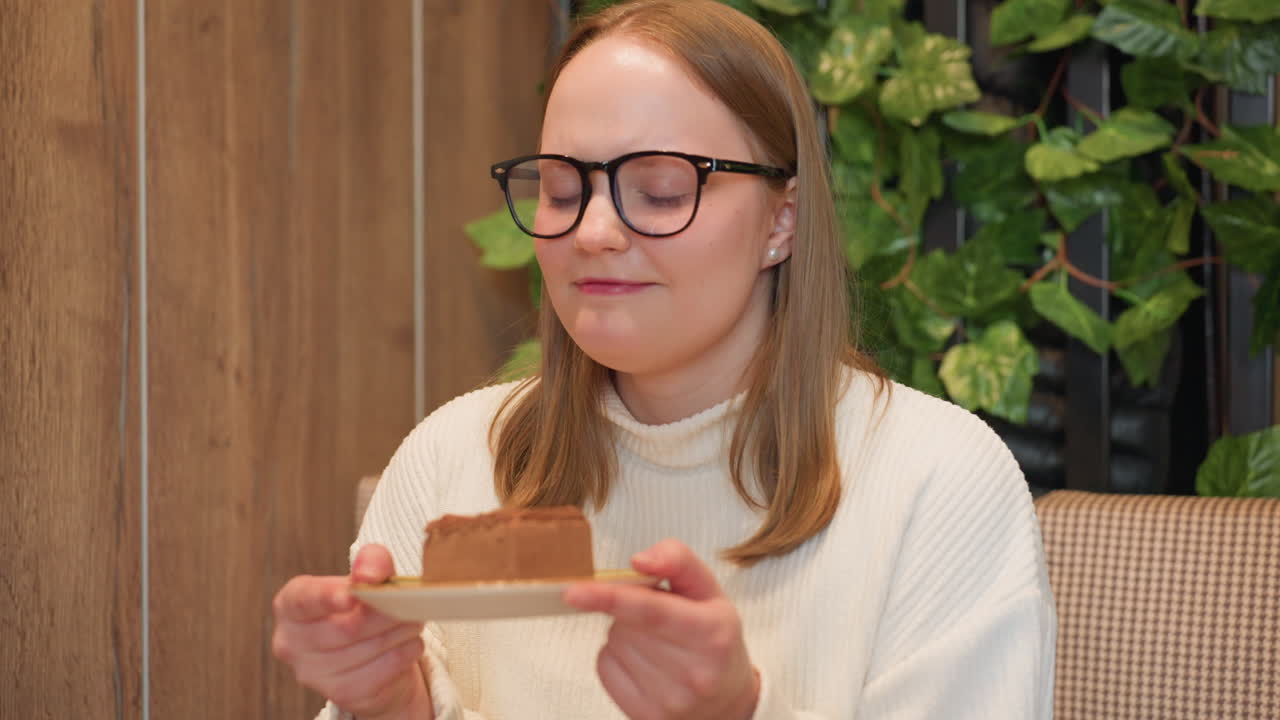 Young woman in glasses and white sweater holds dessert plate near face, smiling warmly as she enjoys aroma of chocolate cake in cozy cafe with wooden walls and lush green backdrop