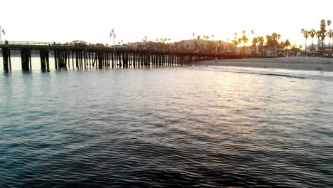 aerial drone shot low over the pacific ocean at sunset off the coast of santa barbara with stearns wharf pier and palm trees in california