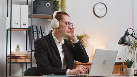 smiling young businessman in headphones listening online music using laptop at home office desk