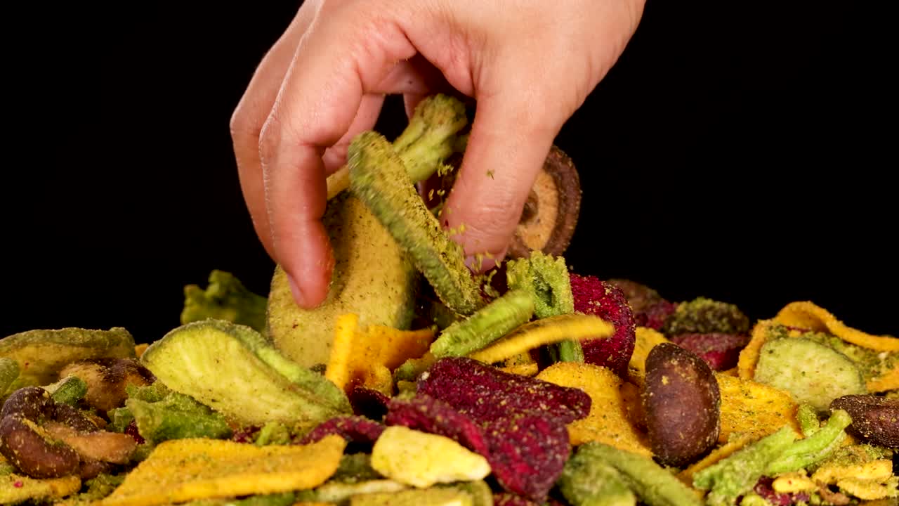 Human hand picks assorted dried vegetable chips under studio lighting, close-up, black background, smooth motion