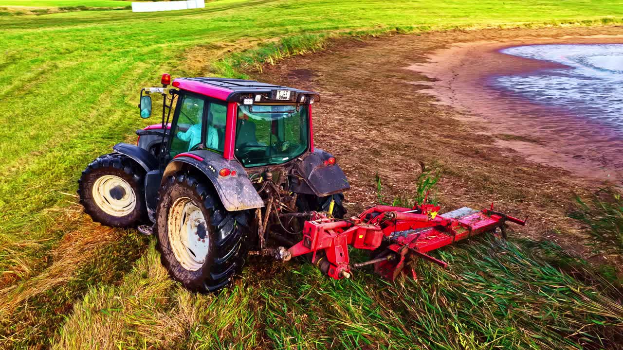 Red tractor mowing grass in green field under bright sky, daytime farming