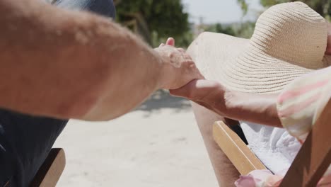 midsection of senior caucasian couple sitting on chairs on sunny beach holding hands, in slow motion