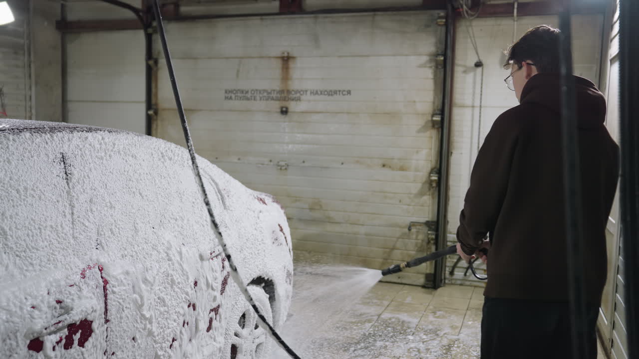 Side view of car washer wearing hoodie spraying thick foam on red vehicle using hose inside industrial warehouse with concrete walls and metallic doors creating authentic working environment