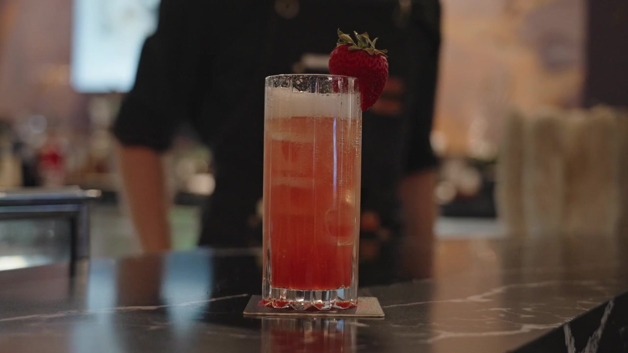 4K-optagelse af en elegant hotelbartender, der skubber en høj lyserød cocktail pyntet med et friskt jordbær ind i fokus, hvilket fremhæver luksuriøs barservice og raffineret præsentation
