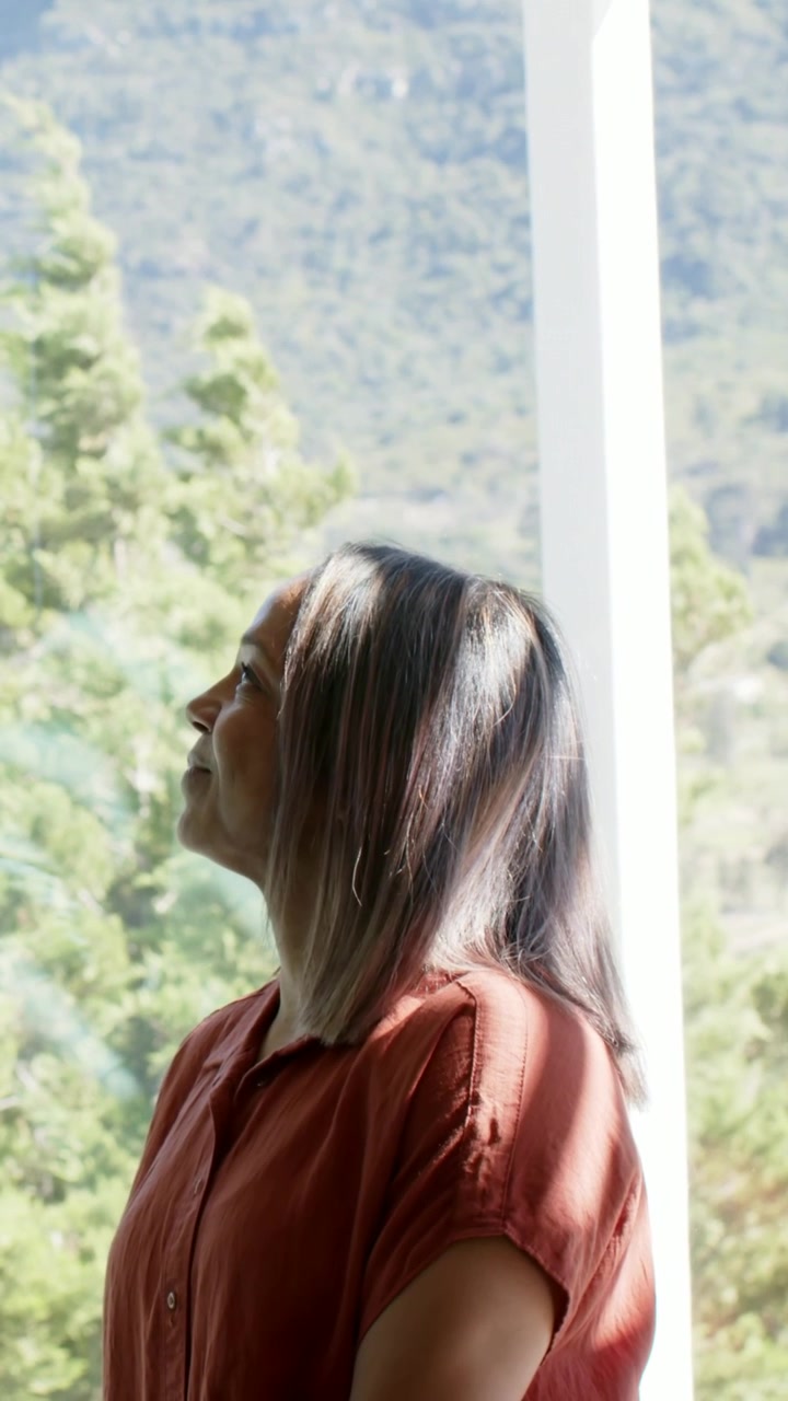 Video verticale: Donna con camicia color ruggine che alza il mento guardando le colline e osserva gli alberi attraverso il vetro di casa.