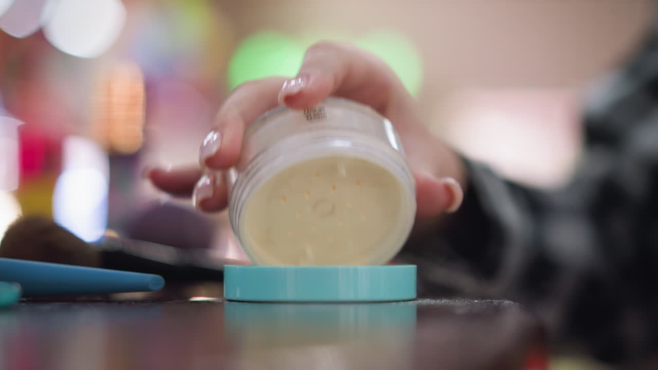 Close-up view of lady hand pouring powder into cover, preparing for makeup application, the cosmetic powder is placed on the table with other beauty tools and products in the background