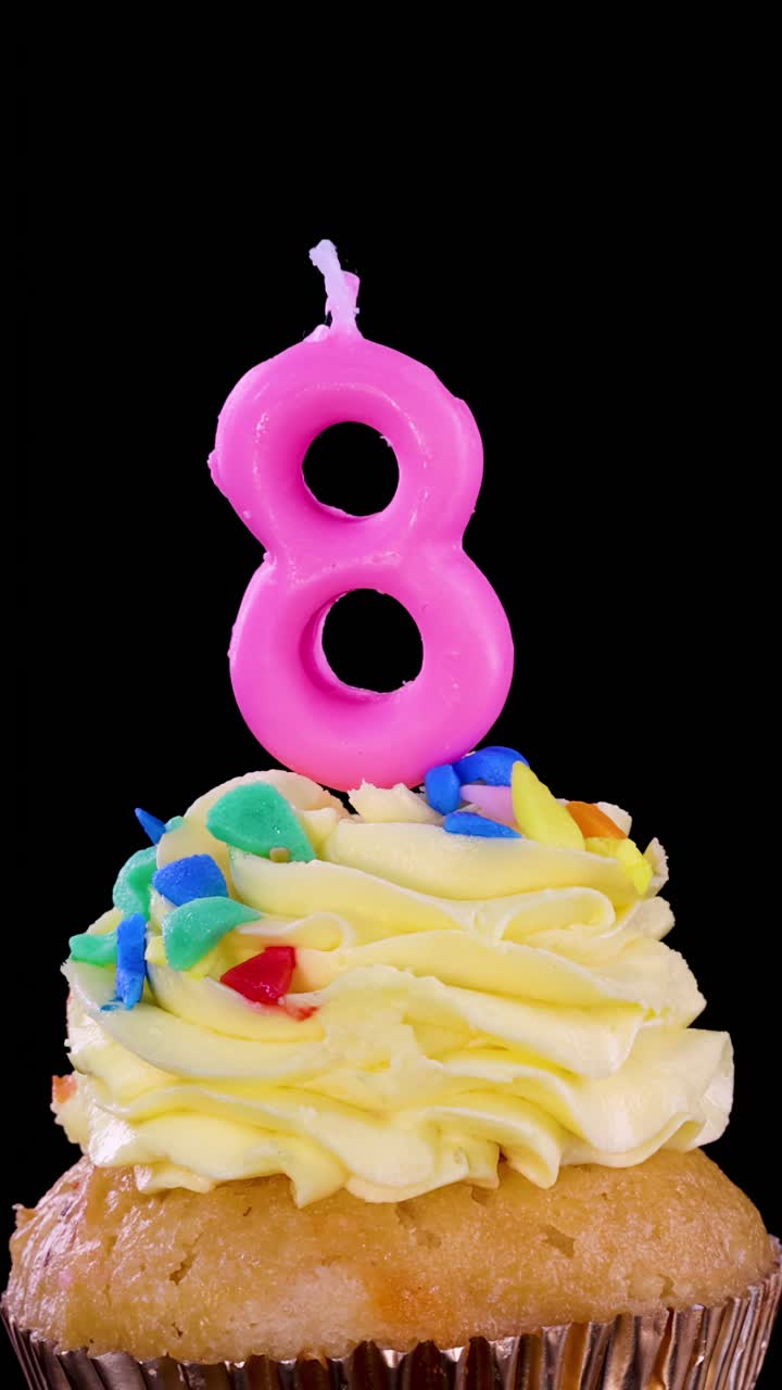 Una mano inserisce una candela rosa a forma di numero otto in un cupcake giallo glassato con codette colorate, su sfondo nero, sotto una luminosa illuminazione da studio