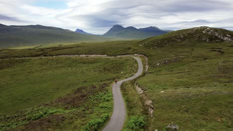 male on longboard riding downhill on a gray asphalt road between the mountains of the scottish wilderness on a cloudy day