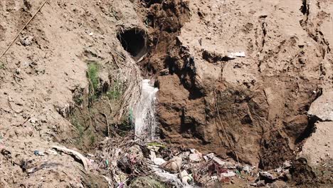 dirty water pouring from hidden sewage pipe into polluted ground filled with trash, urban pollution, health hazard and ecological damage.