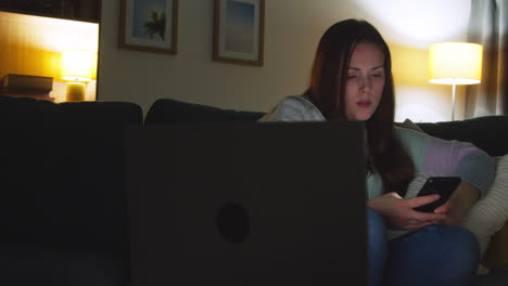 Mujer-Sentada-En-Un-Sofá-En-Casa-Por-La-Noche-Transmitiendo-O-Viendo-Películas-O-Programas-En-Una-Computadora-Portátil-Y-Navegando-Por-Internet-En-El-Teléfono-7