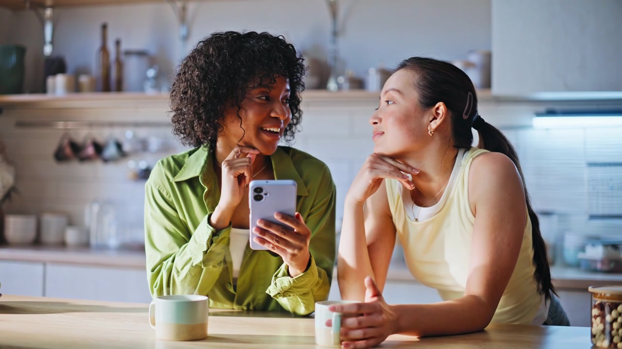 Women browsing phone together kitchen. Friends sharing morning coffee chatting.