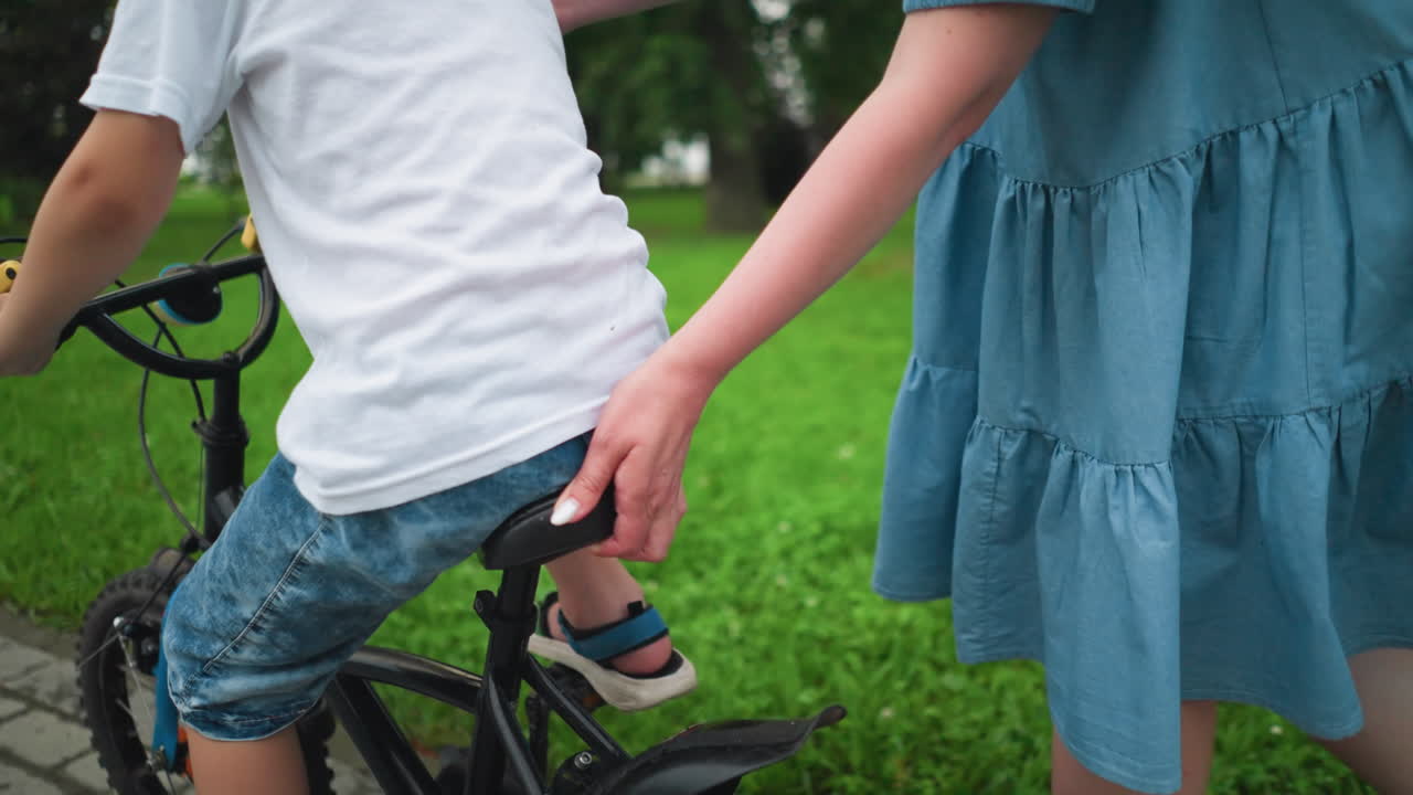 한 여자 는 아들 의 자전거 좌석 의 뒷부분 을 들고, 그 아들 이 초록색 들판 근처 의 산책로 를 따라 달리는 동안 그 아들 을 인도 하고 지원 하고 있다