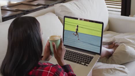 Composite-of-woman-sitting-at-home-holding-coffee-watching-athletics-running-event-on-laptop