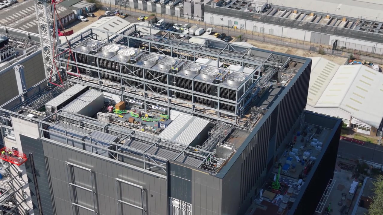 Et luftfoto af et stort datacenter viser dets omfattende kølesystemer på taget. Byggeudstyr antyder igangværende arbejde i denne industripark.