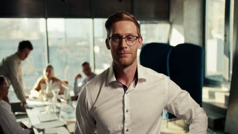 a confident blond man in glasses and a white shirt poses in the office against the background of his gollegs who work at a table in an office with panoramic windows. video filmed in high quality