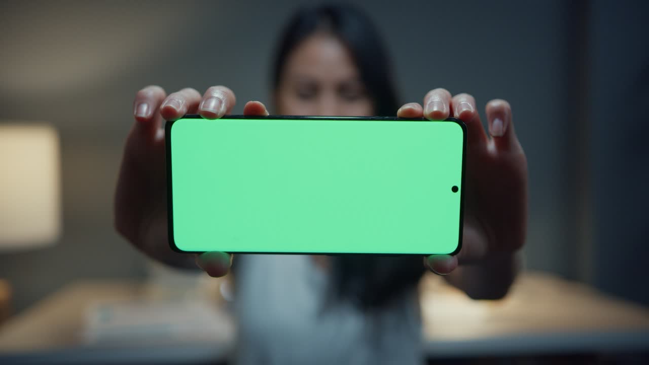 asia joven vestimenta casual inteligente mirando a la cámara, sonreír, feliz y mostrar pantalla simulada teléfono inteligente en la noche de la casa moderna. trabajo desde casa, distancia social, teléfono móvil clave de croma, navegación por internet.