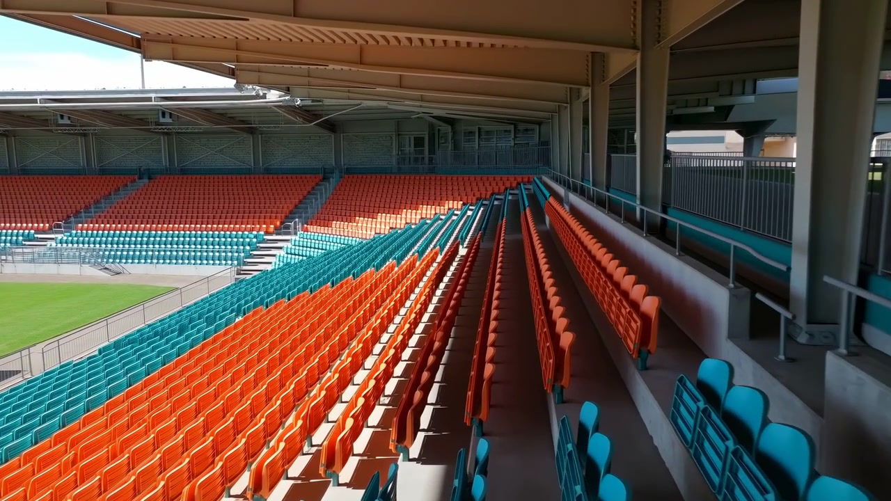 Förhöjd vy från bakre högra gången av en tom modern stadionläktare med omväxlande orange och turkosa plastsäten under ett metalltak. Stålpelare.