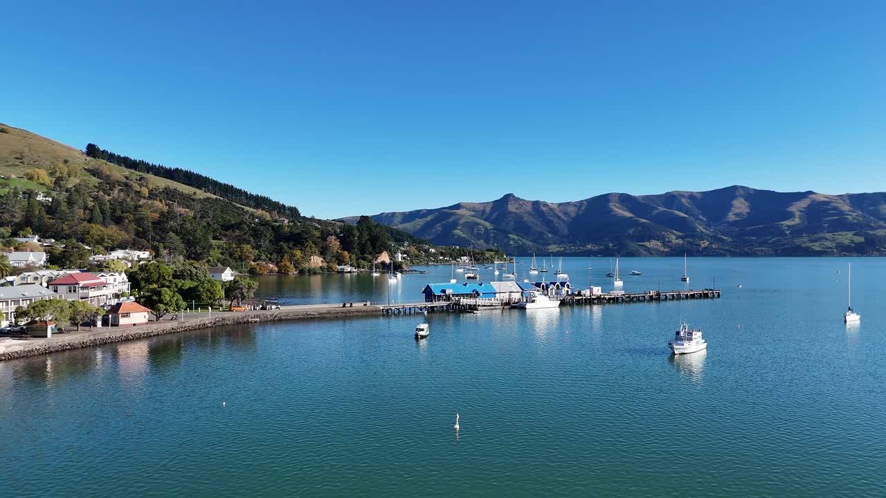 Drone footage captures serene Akaroa Bay, showcasing boats, calm waters, and surrounding hills under clear blue skies