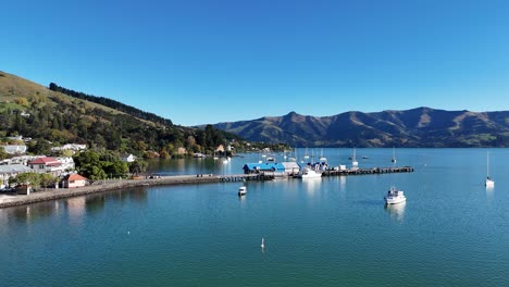 drone footage captures serene akaroa bay, showcasing boats, calm waters, and surrounding hills under clear blue skies
