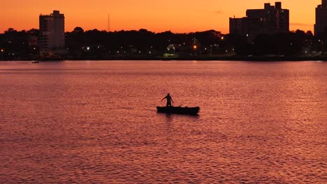 Foto-Panorámica-De-Un-Pescador-En-Canoa,-Navegando-Por-El-Río-Paraná-Con-La-Ciudad-De-Posadas-Al-Atardecer-Al-Fondo,-Argentina.