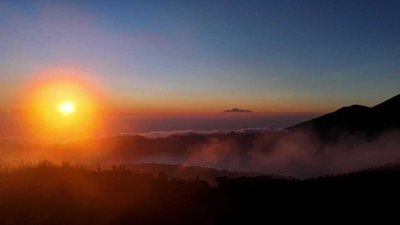 A golden sunrise cast light over Mount Batur in Bali, Indonesia, with hikers gathered at the summit to witness the mist veiling volcanic peak and glowing clouds