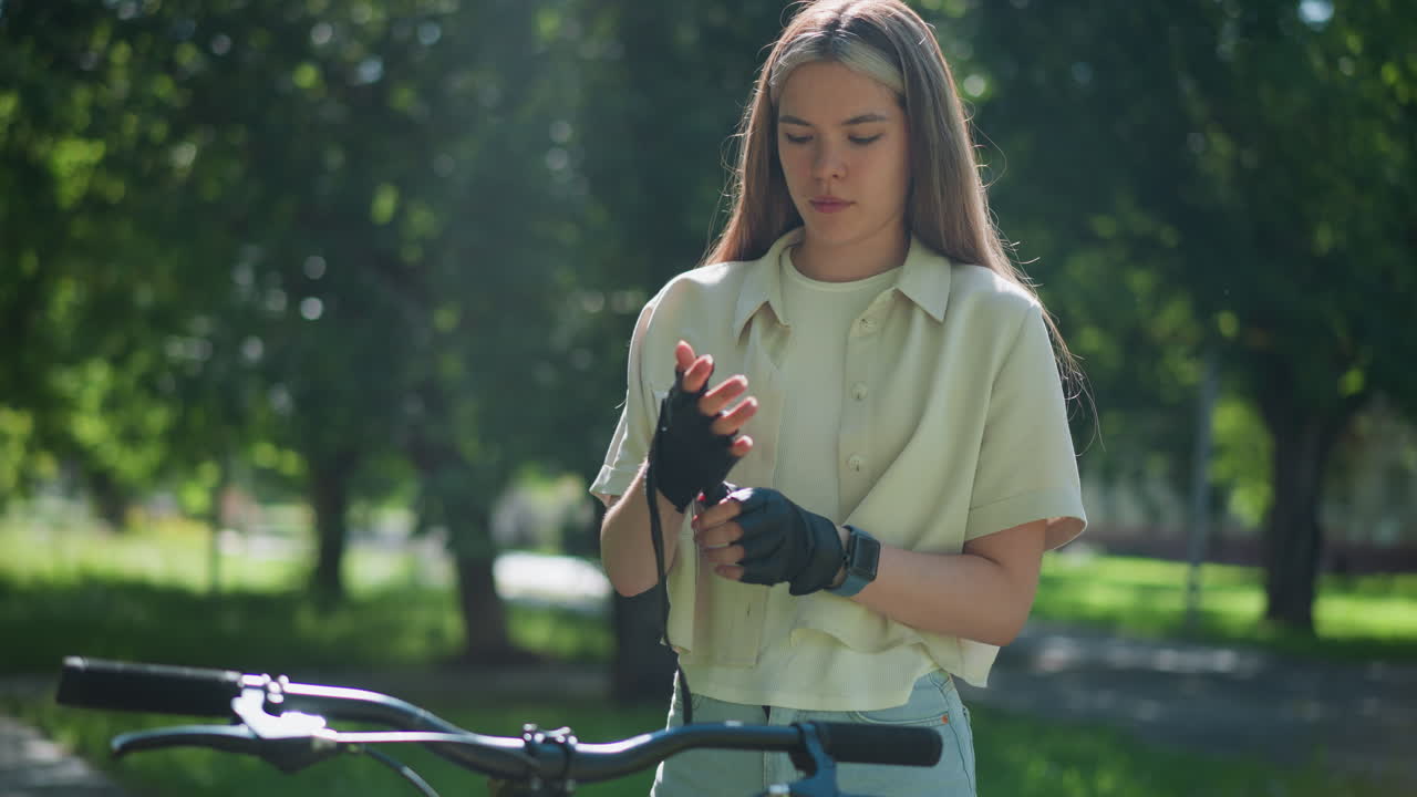 una mujer joven está de pie al lado de su bicicleta, ajustando cuidadosamente el guante de motociclista en su mano derecha, la expresión enfocada en su rostro, la luz del sol se filtra a través de los árboles