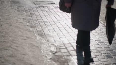 leg level view shows people in sturdy boots walking along icy pavement, long shadows stretching over wet bricks, winter light glinting off slush and snow, city traffic moving past curb in morning