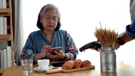 Active-senior-Asian-woman-working-in-a-coffeeshop-using-a-mobile-device,-smartwatch-for-NFC,-QR-code-scanning,-contactless-payment-to-pay-for-coffee-and-snack