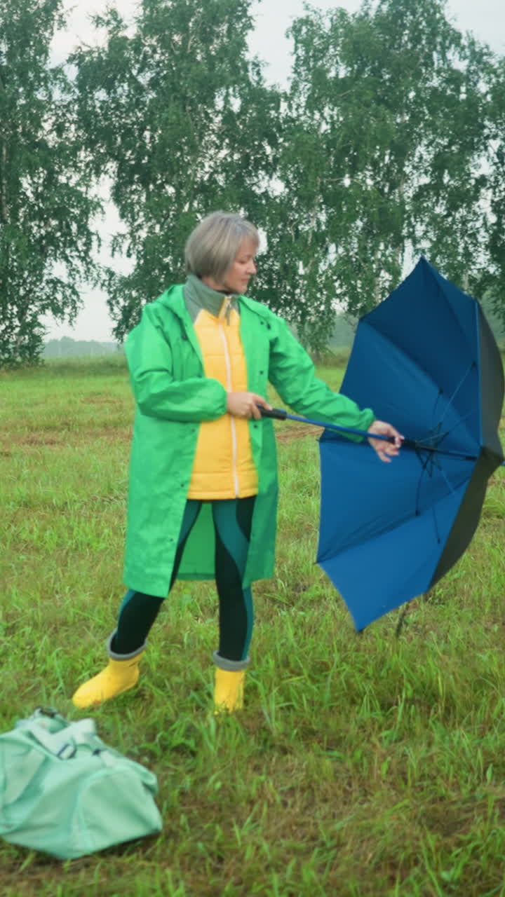 초록색 레인코트를 입은 중년 여성이 잔디에 서서 우산을 닫고 바닥에 있는 가방 근처에 떨어뜨립니다.