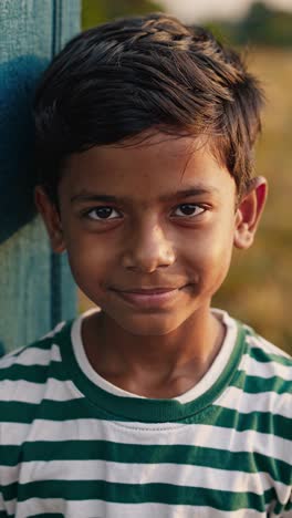 portrait of a smiling boy wearing a striped t shirt, leaning against a wooden fence in a field, bathed in the warm light of the golden hour