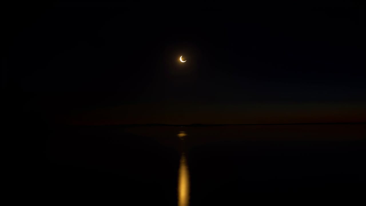 Una falce di luna luminosa pende sull'acqua immobile mentre il suo caldo riflesso verticale tremola dall'orizzonte lontano verso il primo piano, incorniciata da un cielo scuro e una debole fascia arancione di crepuscolo