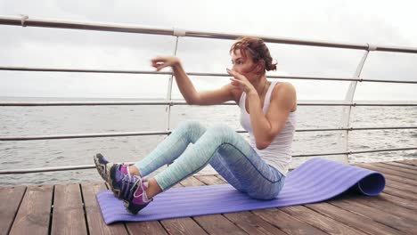 Young-athletic-woman-doing-situps-on-the-mat-and-boxing-doing-kicks-by-the-ocean-in-the-early-morning.-Fitness-woman-doing-abs