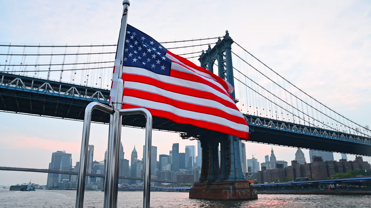 City skyline with flag under bridge. An American flag waves on a boat while passing under the Manhattan Bridge at sunset, with a skyline backdrop